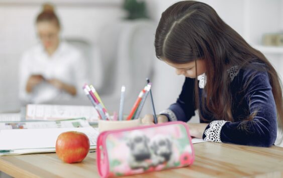 Focused young girl in school uniform sitting at table with stationery and apple while writing in copybook during studies against mother in modern apartment