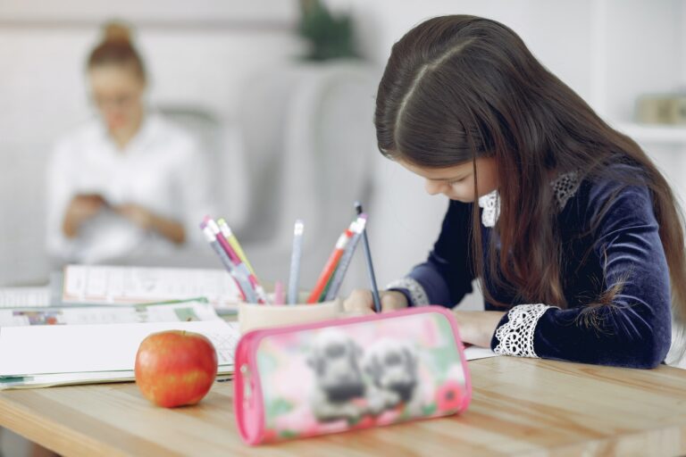 Focused young girl in school uniform sitting at table with stationery and apple while writing in copybook during studies against mother in modern apartment